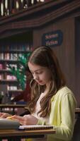 Vertical Focused schoolgirl reading a book in the library for mandatory literature, gaining knowledge for her academic curriculum during study time. Young scholar focusing on lecture tasks. Camera A. video