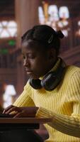 Vertical African american schoolgirl writing class notes on laptop in a library, working on a project with online information at a study desk. Pupil preparing for lessons at the academic institute. Camera A. video