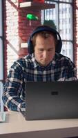 Teleworker enjoying music while researching data for company project, doing tasks in personal home office. Man sitting at desk in front of laptop and using headphones, camera A video