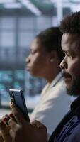 Vertical Worker in data center wearing headphones taking break from implementing AI systems, using smartphone. Focus on artificial intelligence server farm employee using mobile phone, camera A video