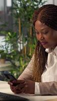 Vertical African american manager takes a break from business work and browsing social media, checks messages and websites at her desk. Analyst multitasking and texting during schedule. Camera A. video