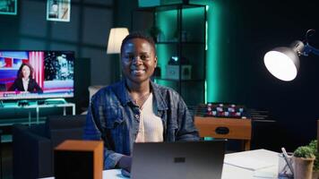 Portrait of jolly woman sitting on chair at home office desk in neon illuminated apartment late at night. Cheerful person in RGB lit living room enjoying leisure time, camera B video