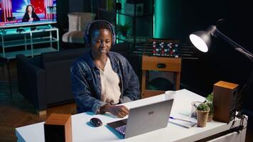 Freelancer doing computer tasks for team project in personal home office while listening music. Remote employee working late at night, typing on laptop keyboard, putting headphones on, camera B video