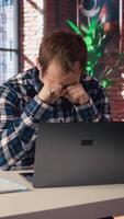 Tired man in living room office shrugging head, upset after having to answer too many emails on laptop. Exhausted freelancer feeling overworked while typing on device keyboard, camera A video