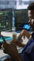 Vertical Admin in server room using mobile phone, relaxing after ensuring minimal downtime. Data center IT expert texting on smartphone, taking break from monitoring performance, camera A video