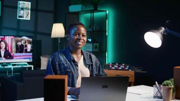 Portrait of happy woman sitting at home office desk, preparing to solve tasks on laptop. Smiling freelancer in front of digital device, doing email communication in living room, camera B video