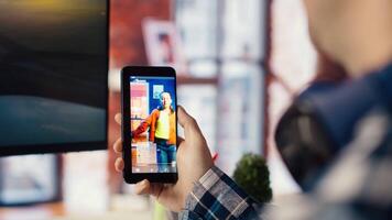 Man in workspace watching videos on social media on smartphone. Person using social network app on mobile phone with computer monitor idle in front of him showing screensaver, camera B