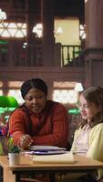Vertical African american teacher providing instruction to diverse girls during private sessions, assisting with homework and school projects in a library. Doing tutoring activity for success. Camera A. video