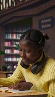 Vertical Young focused scholar doing her homework at a library desk after classes, engrossed in literature and notes. Pupil completing her notes in the copybook, productive study sessions. Camera A. video