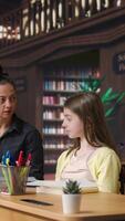 Vertical Caucasian tutor guiding elementary school girls at a library desk, providing private lessons to aid in academic development with work books and notes. Teacher discussing with scholars. Camera A. video