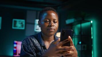 Smiling woman at home texting friend on cellphone, taking break from remote job in personal office. Upbeat freelancer relaxing in apartment, talking with mate on internet using phone app, camera B video