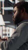Vertical Black male CEO dealing with back pain and stretching his muscles at the desk, sitting with an incorrect posture and trying to fix it. General manager working with data analytics. Camera A. video