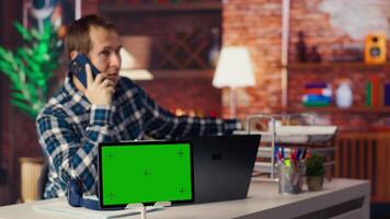 Green screen tablet next to man using laptop, answering phone call while checking emails displayed on screen. Mockup device next to worker in telephone call with friend at work, camera B video