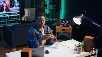 African american woman relaxing in apartment, texting friends on cellphone, enjoying leisure time. Smiling remote worker taking break to talk with mates on internet using phone, camera B video