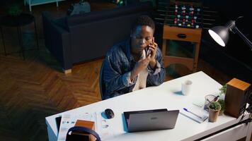 African american teleworker receiving phone call from job colleague while working from home on laptop. Freelancer speaking with coworker using smartphone while solving tasks, camera B video