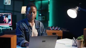 Porträt von lächelnd afrikanisch amerikanisch Frau beim Schreibtisch Tippen auf Laptop Klaviatur, lösen Aufgaben. froh Fernbedienung Arbeiter mit Notizbuch zu senden E-Mails zu Geschäft Partner von Leben Zimmer, Kamera b video