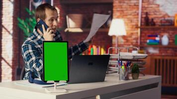 Chroma key tablet next to freelancer in home office sitting at desk in front of laptop, answering phone call. Isolated screen device next to freelancer receiving telephone call on smartphone, camera B video