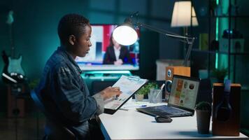 African american woman working from home, using laptop, looking at statistic charts. Remote worker in personal apartment office crosschecking important info between clipboard and notebook, camera A video