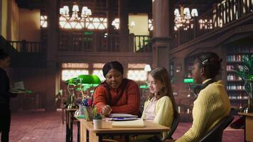 African american teacher providing instruction to diverse girls during private sessions, assisting with homework and school projects in a library. Doing tutoring activity for success. Camera A. video