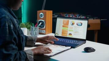 empreendedor trabalhando a partir de lar, usando computador portátil, escrevendo notas em papel. controlo remoto trabalhador dentro pessoal apartamento escritório transcrever importante informação em bloco de anotações, olhando às estatística gráficos, Câmera uma video