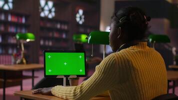 Black scholar does research via internet and taking notes next to chroma key in a library, concentrating on theoretical studies and completing her class notes for the school assignment. Camera B. video