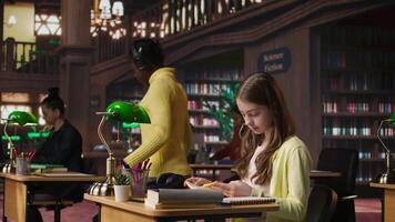 Focused schoolgirl reading a book in the library for mandatory literature, gaining knowledge for her academic curriculum during study time. Young scholar focusing on lecture tasks. Camera A. video