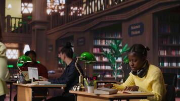 African american teen girl working hard on developing writing skills at the library, trying to create an essay with information from a book. Completing school assignment with notebooks. Camera B. video