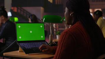 Black confident librarian typing on laptop with green screen in a quiet library workspace, organizing resources and mentoring students in their academic journey with pride and expertise. Camera B. video
