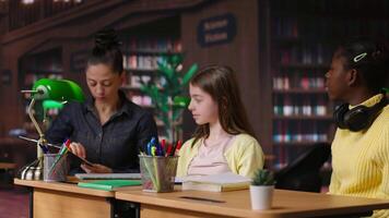 Caucasian tutor guiding elementary school girls at a library desk, providing private lessons to aid in academic development with work books and notes. Teacher discussing with scholars. Camera B. video