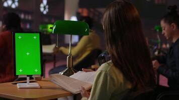 Schoolgirl reading a book next to copy space on tablet at the library desk, using academic database for research. Completing school assignments and developing her educational skills. Camera B. video