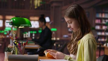 Focused schoolgirl reading a book in the library for mandatory literature, gaining knowledge for her academic curriculum during study time. Young scholar focusing on lecture tasks. Camera B. video