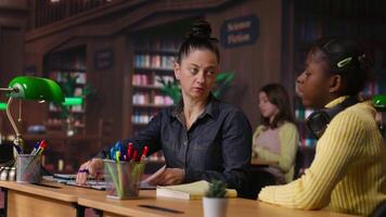 Caucasian tutor guiding elementary school girl at a library desk, providing private lessons to aid in their academic development with work books and notes. Teacher discussing with scholar. Camera B. video