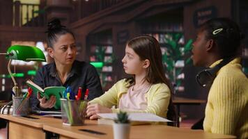 Female tutor mentoring schoolgirls in a school library setting, boosting their understanding of a subject through personalized guidance. Teacher doing private lessons with two pupils. Camera B. video
