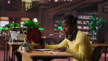 Young focused scholar doing her homework at a library desk after classes, engrossed in literature and notes. Pupil completing her notes in the copybook, productive study sessions. Camera B. video