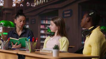 Elementary students receiving support from a tutor working on homework assignments in a diverse learning environment in the school library. Professor guiding her pupils before class. Camera B. video