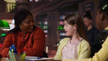 African american private tutor offering support to diverse schoolgirls, guiding them through a lesson plan at a library desk. Teacher providing learning materials, mentoring scholars. Camera B. video