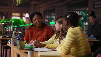 Black teacher mentoring her teen girls students with an assignment, teaching academic concepts and enhancing their knowledge through practical exercises. Private lessons at the library. Camera B. video