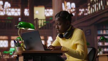 African american schoolgirl writing class notes on laptop in a library, working on a project with online information at a study desk. Pupil preparing for lessons at the academic institute. Camera A. video