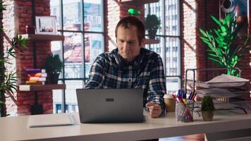 Freelancer in home office sitting at desk, excited after having success with project online, looking at laptop screen. Man celebrating after receiving good news, looking at notebook screen, camera A video