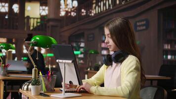 Preteen student using a tablet to complete her class notes in the library, uses the digital academic database to consult learning materials. Preparing diligently to pass her exams. Camera A. video