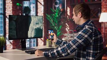Man in workspace watching videos on social media on smartphone. Person using social network app on mobile phone with computer monitor idle in front of him showing screensaver, camera A