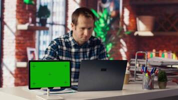 Green screen tablet next to employee completing spreadsheets on laptop and listening music. Chroma key device next to worker in home office wearing headphones editing documents, camera B video