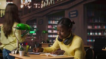 Young focused scholar doing her homework at a library desk after classes, engrossed in literature and notes. Pupil completing her notes in the copybook, productive study sessions. Camera A. video
