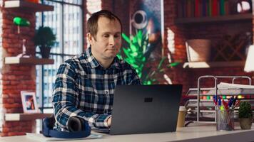 Man seated at home office desk using laptop, answering phone call while checking emails displayed on screen. Worker in telephone call with friend at work while reading messages in inbox, camera B video
