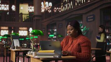 Happy female librarian typing on her laptop in a quiet library workspace, dedicated to organizing resources and mentoring students in their academic journey with pride and expertise. Camera A. video