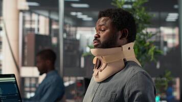 Hurt employee with a cervical collar working on a new business project, struggling to sit at the desk with his neck fracture. African american company associate tries to solve tasks. Camera A. video