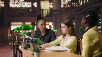 Female tutor mentoring schoolgirls in a school library setting, boosting their understanding of a subject through personalized guidance. Teacher doing private lessons with two pupils. Camera A. video