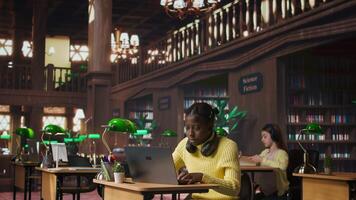 African american schoolgirl writing class notes on laptop in a library, working on a project with online information at a study desk. Pupil preparing for lessons at the academic institute. Camera B. video