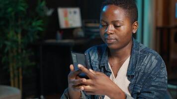 Smiling woman at home texting friend on cellphone, taking break from remote job in personal office. Upbeat freelancer relaxing in apartment, talking with mate on internet using phone app, camera A video