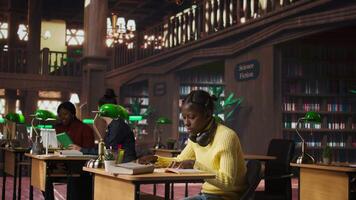 African american teen girl working hard on developing writing skills at the library, trying to create an essay with information from a book. Completing school assignment with notebooks. Camera A. video